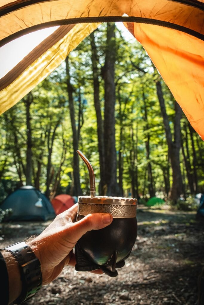 Mano sosteniendo una taza de mate en medio de un campamento rodeado de árboles.