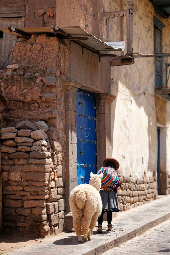 Mujer andina con vestimenta tradicional y alpaca frente a una antigua puerta azul en calle empedrada de Cusco
