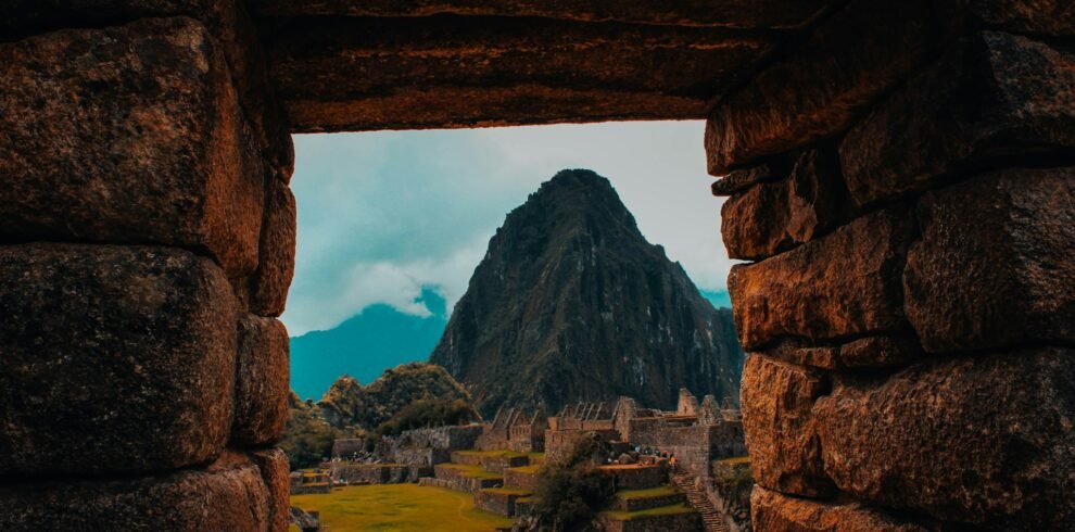 Vista de Machu Picchu y Huayna Picchu enmarcada por ventana de piedra inca, mostrando la ciudadela, terrazas verdes y montaña icónica bajo cielo azul nublado
