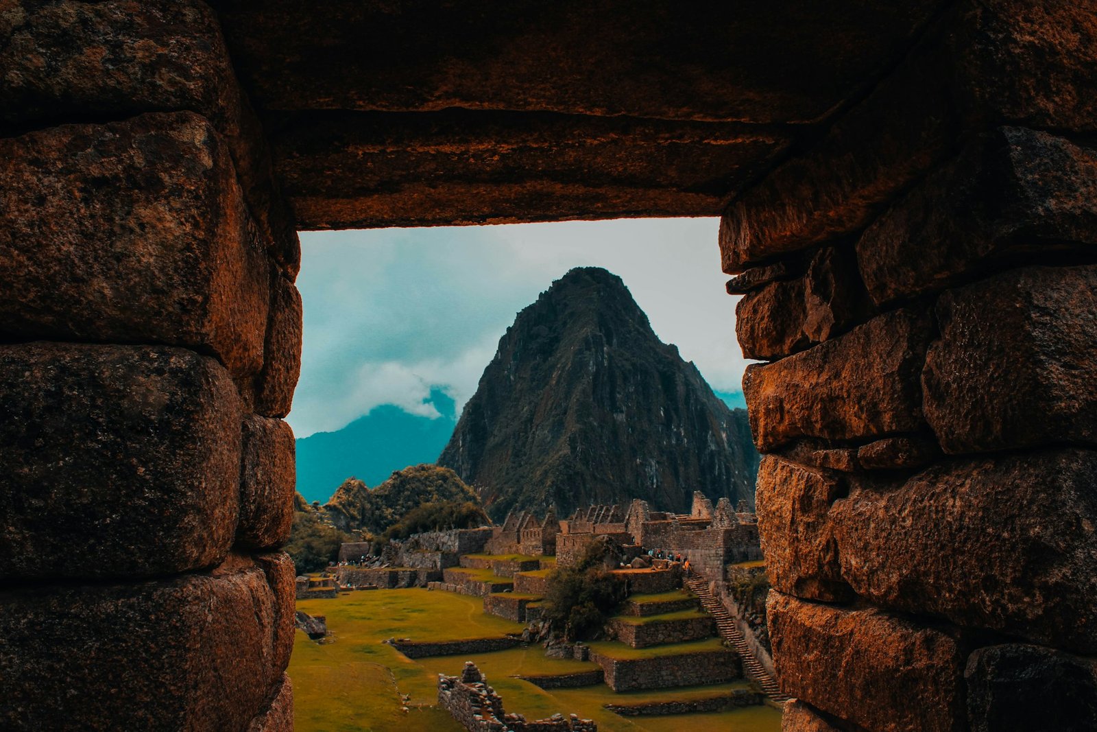 Vista de Machu Picchu y Huayna Picchu enmarcada por ventana de piedra inca, mostrando la ciudadela, terrazas verdes y montaña icónica bajo cielo azul nublado
