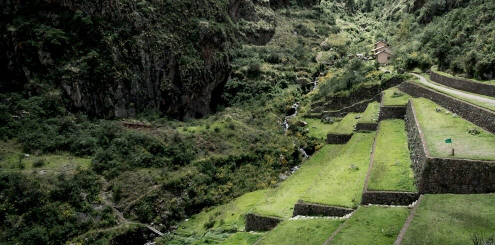 Terrazas agrícolas incas con muros de piedra y campos verdes descendiendo hacia un valle con río, rodeadas de montañas escarpadas en el Valle Sagrado, Cusco.