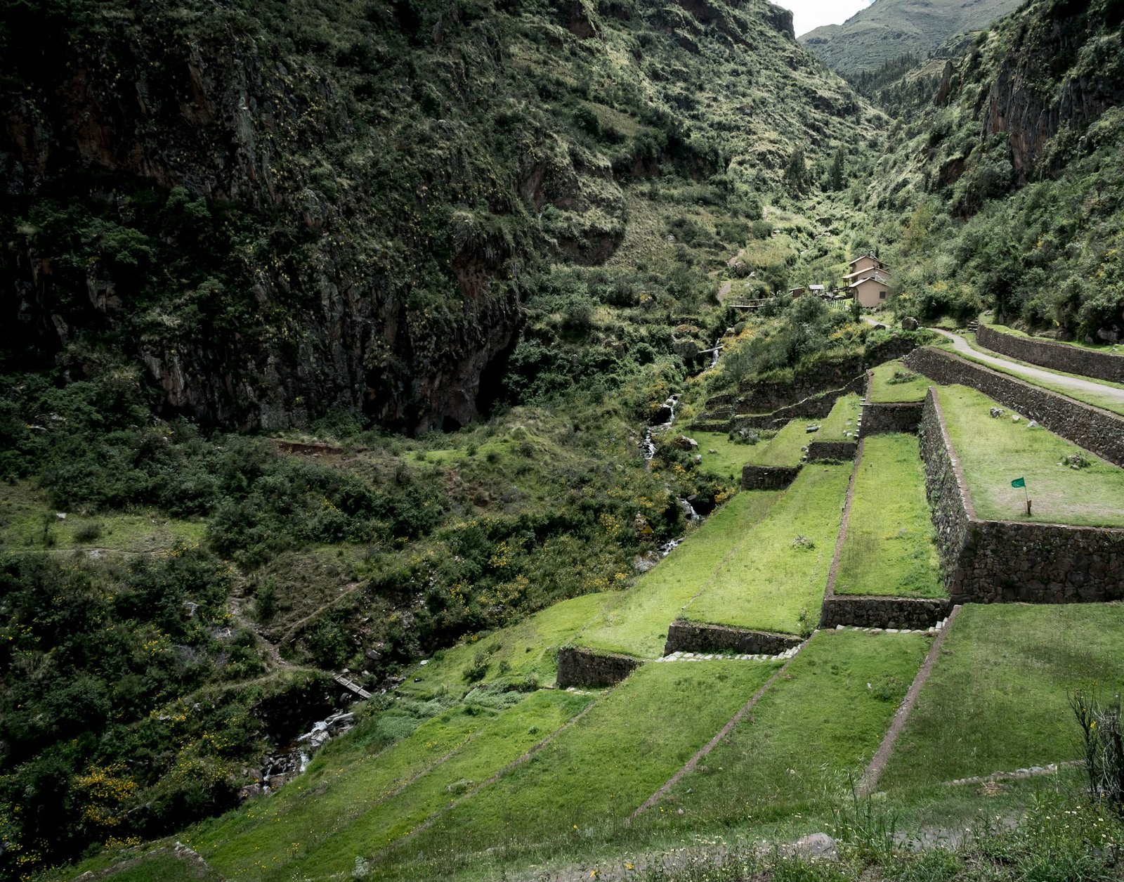 Terrazas agrícolas incas con muros de piedra y campos verdes descendiendo hacia un valle con río, rodeadas de montañas escarpadas en el Valle Sagrado, Cusco.