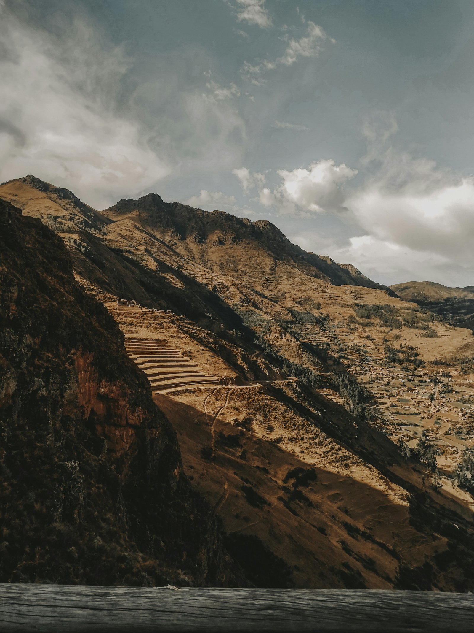 Vista panorámica de las terrazas agrícolas incas en Pisac, mostrando andenes escalonados en la ladera de la montaña con el Valle Sagrado al fondo bajo cielo azul con nubes.